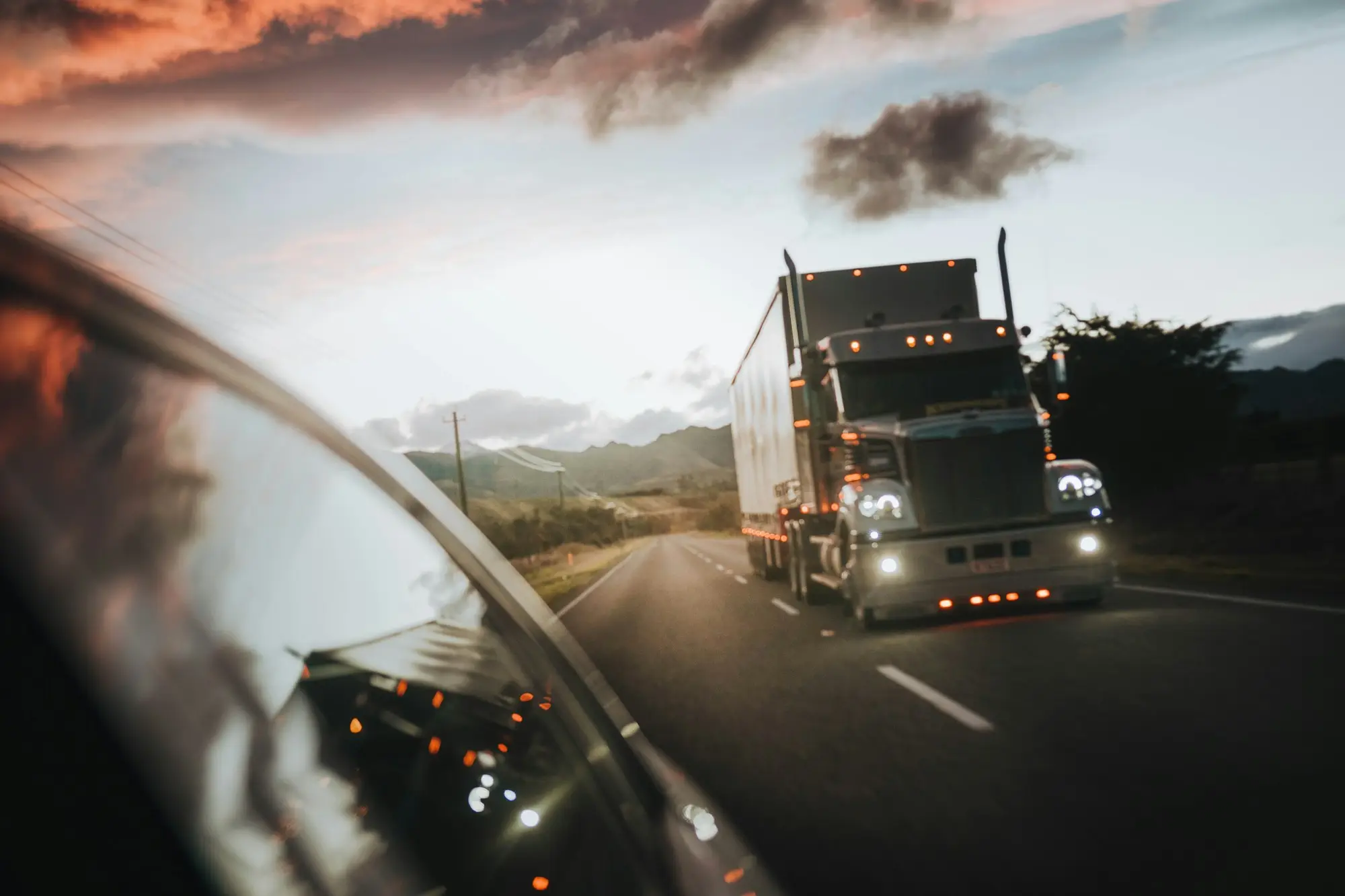 Bonneted prime mover with orange marker lights approaching on a two-lane country highway at dusk, seen past a ute's side mirror