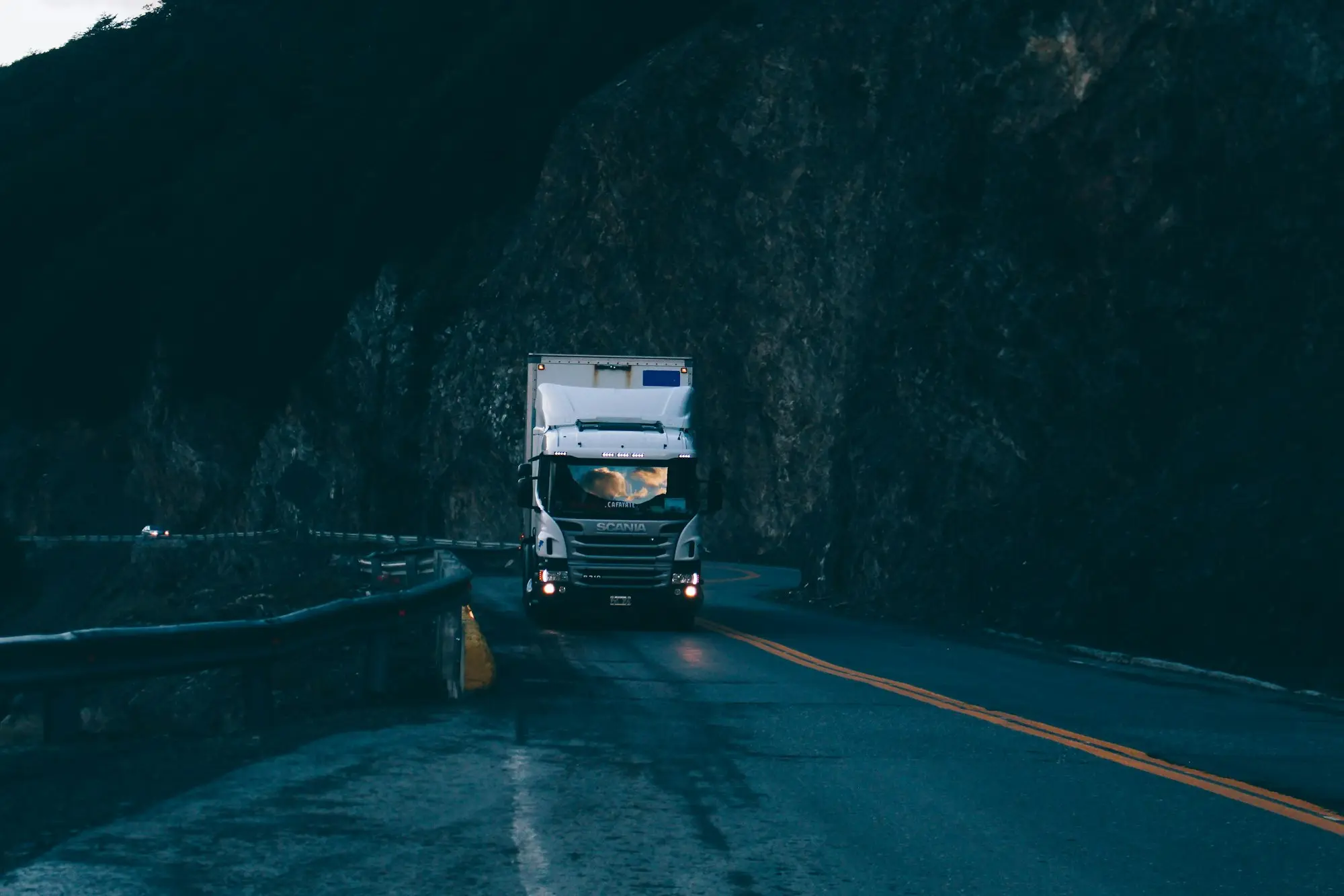 Cab-over prime mover with a box trailer rounding a dark mountain road at dusk, headlights on